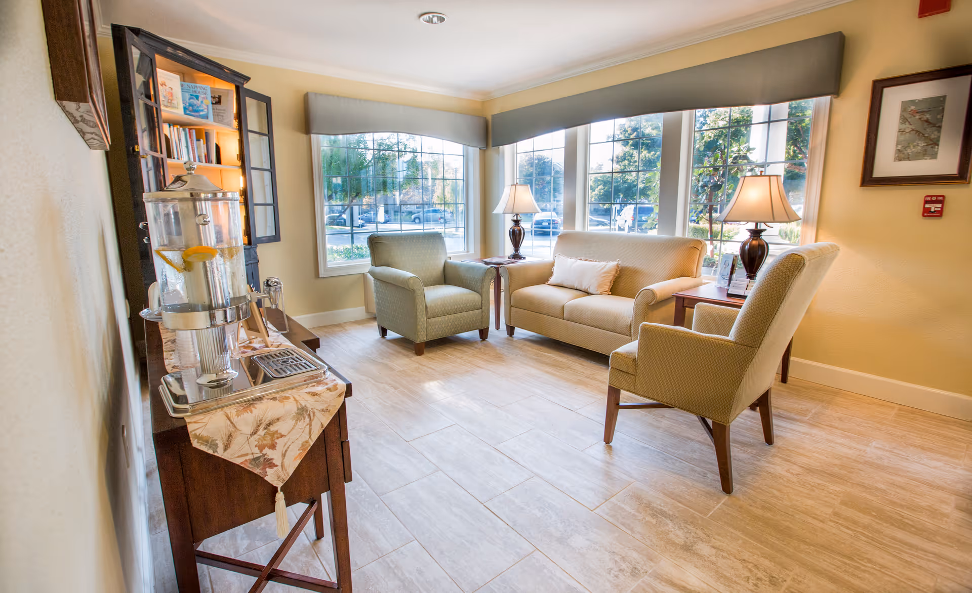 A bright and cozy sitting area with large windows letting in natural light. The room features a beige loveseat with a white pillow, two green upholstered armchairs, two wooden side tables with lamps, and a wooden cabinet with books and a water dispenser on top. The floor is tiled with light-colored tiles, and the walls are painted a soft yellow.