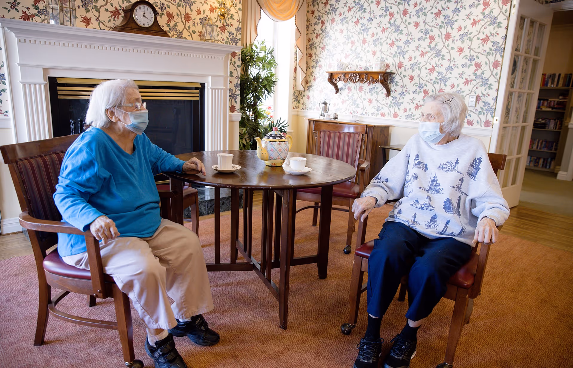Two elderly women wearing face masks sitting on wooden chairs around a round wooden table with teacups in a cozy room with floral wallpaper, a fireplace, and a plant near the window.