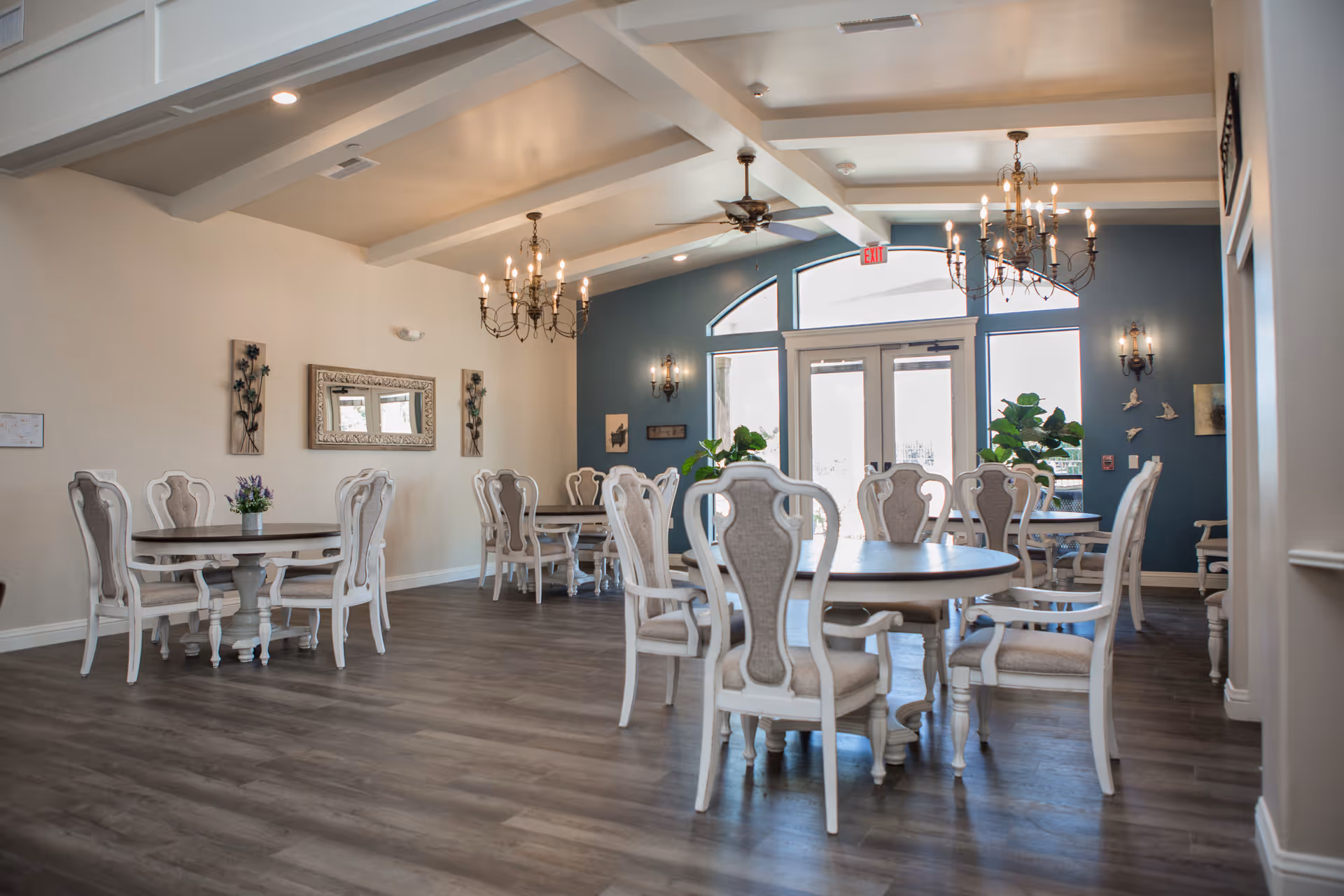 Spacious, well-lit dining room with round tables, upholstered chairs, chandeliers, and large windows/doors.