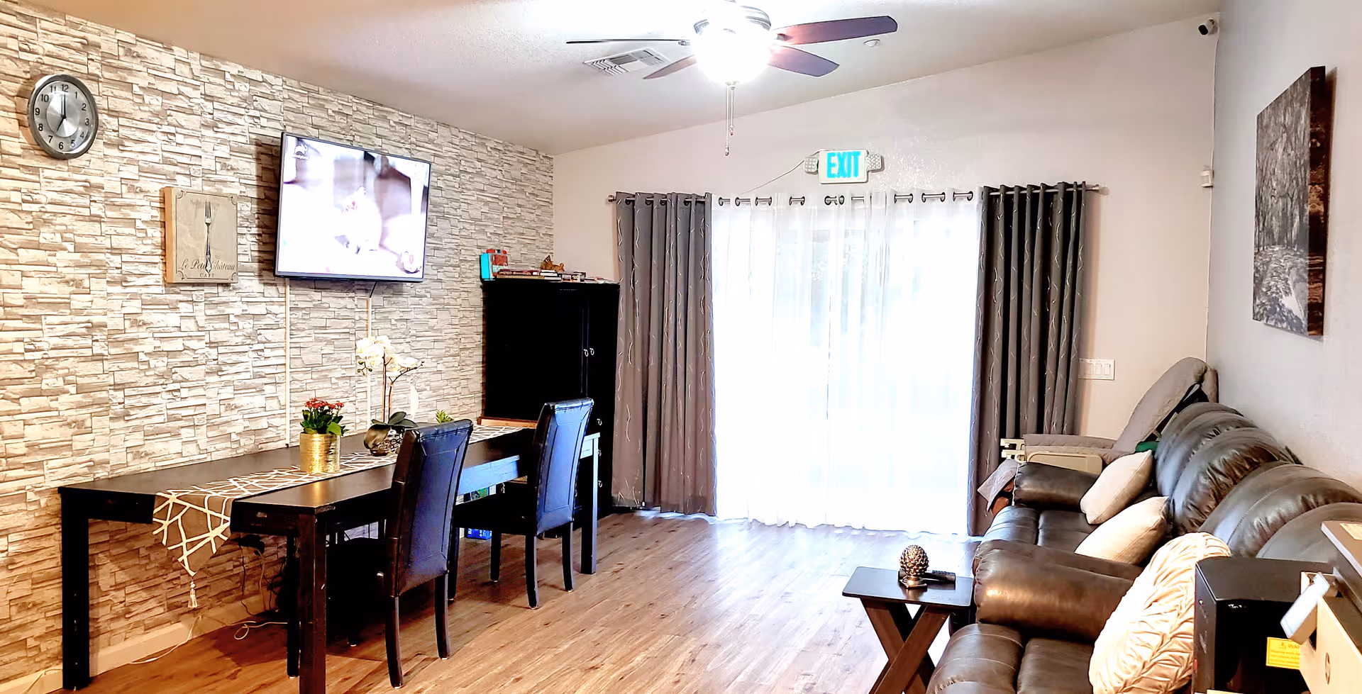 Open living room and dining area with a leather sofa, wall-mounted TV on a stone accent wall, a dining table with chairs, and sliding glass doors with curtains.