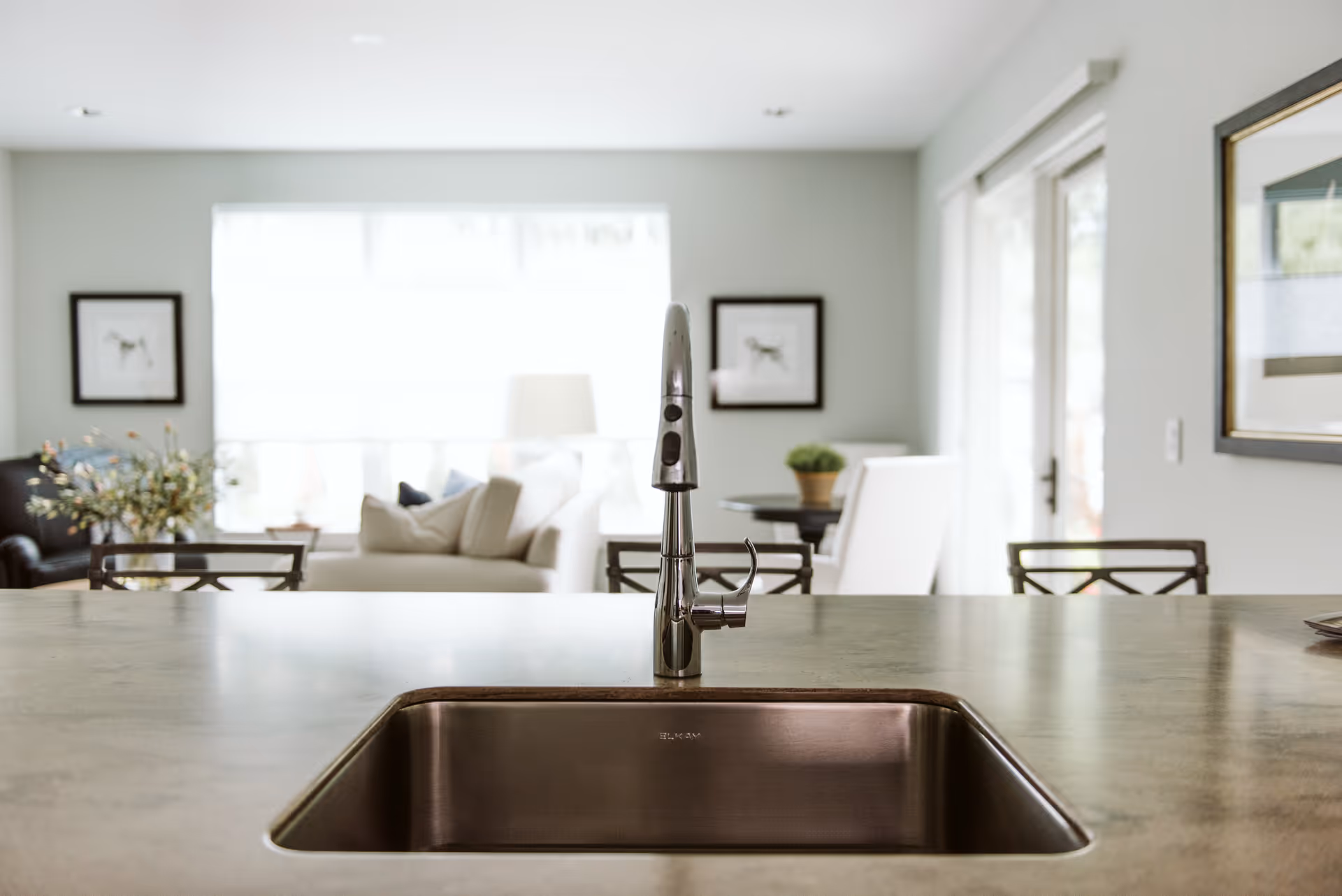 Stainless steel kitchen sink and faucet on an island with a bright open living and dining area visible in the background.