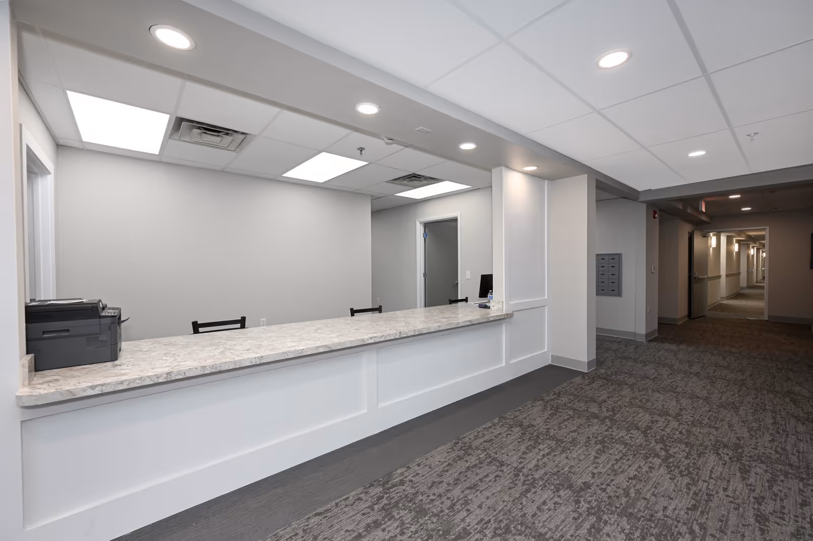 Reception area with a long marble countertop, two black chairs behind the counter, a printer on the left side, and a hallway extending to the right with carpeted floors and white walls under a drop ceiling with recessed lighting.