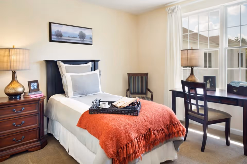 A cozy bedroom with a single bed featuring white and gray bedding and an orange throw blanket. A black tray with a cup and an open book rests on the bed. To the left is a wooden nightstand with a lamp, books, and a framed photo. A wooden chair with striped upholstery is against the wall. On the right side, there is a wooden desk with a lamp, a framed photo, and a box, positioned in front of a large window with white curtains letting in natural light. A landscape painting hangs above the bed.