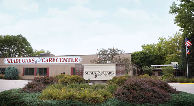 Exterior view of Shady Oaks Care Center building with a sign in front surrounded by bushes and trees. The building is single-story with a beige facade and red window shutters. There are American and other flags on flagpoles to the right side of the image.