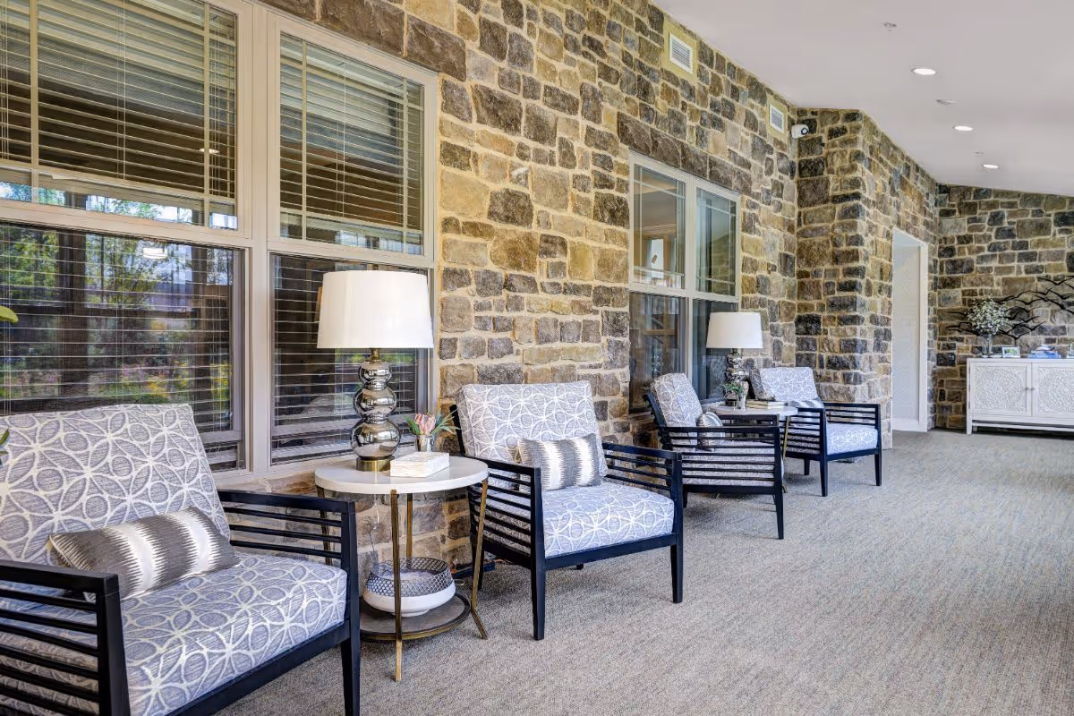 A bright and spacious interior hallway with a stone wall on one side and large windows. The hallway is furnished with several cushioned armchairs with patterned upholstery, each paired with a small round side table holding a lamp and decorative items. The floor is carpeted, and recessed ceiling lights illuminate the space.