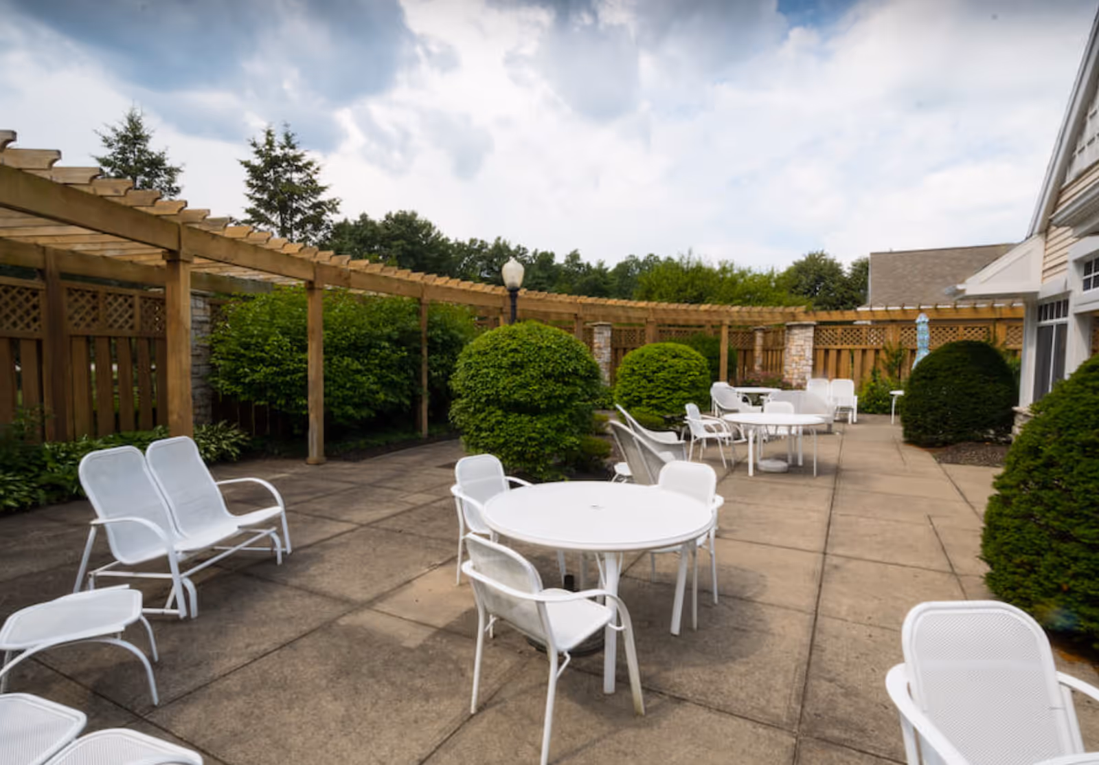 Outdoor patio area with white metal tables and chairs arranged on a concrete surface, surrounded by green bushes and wooden pergola structures under a partly cloudy sky.
