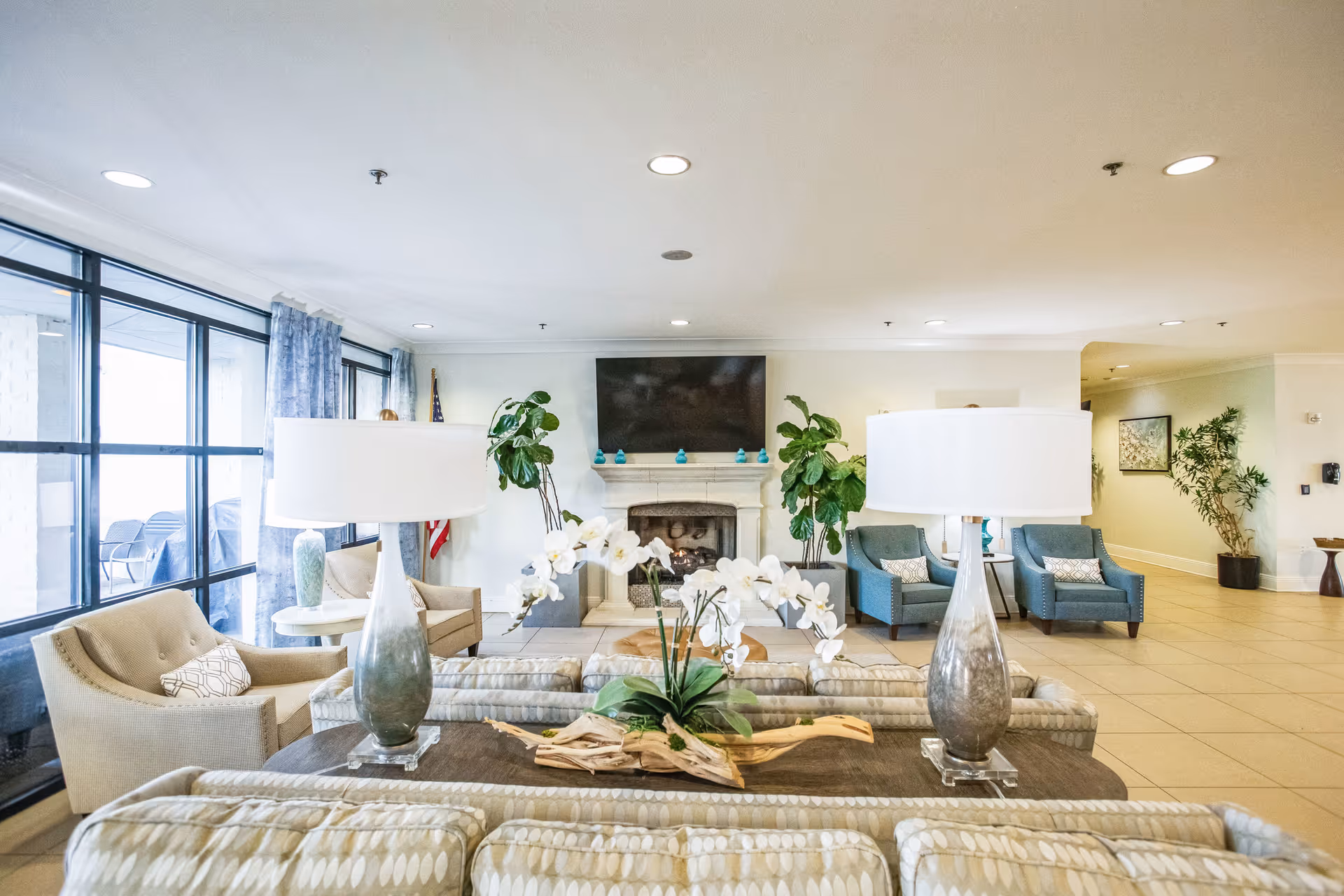 A bright and spacious living room area in The Blake at Township facility featuring patterned sofas, two large table lamps, a coffee table with a floral centerpiece, armchairs, a fireplace with a mounted flat-screen TV above it, and large windows with blue curtains letting in natural light.