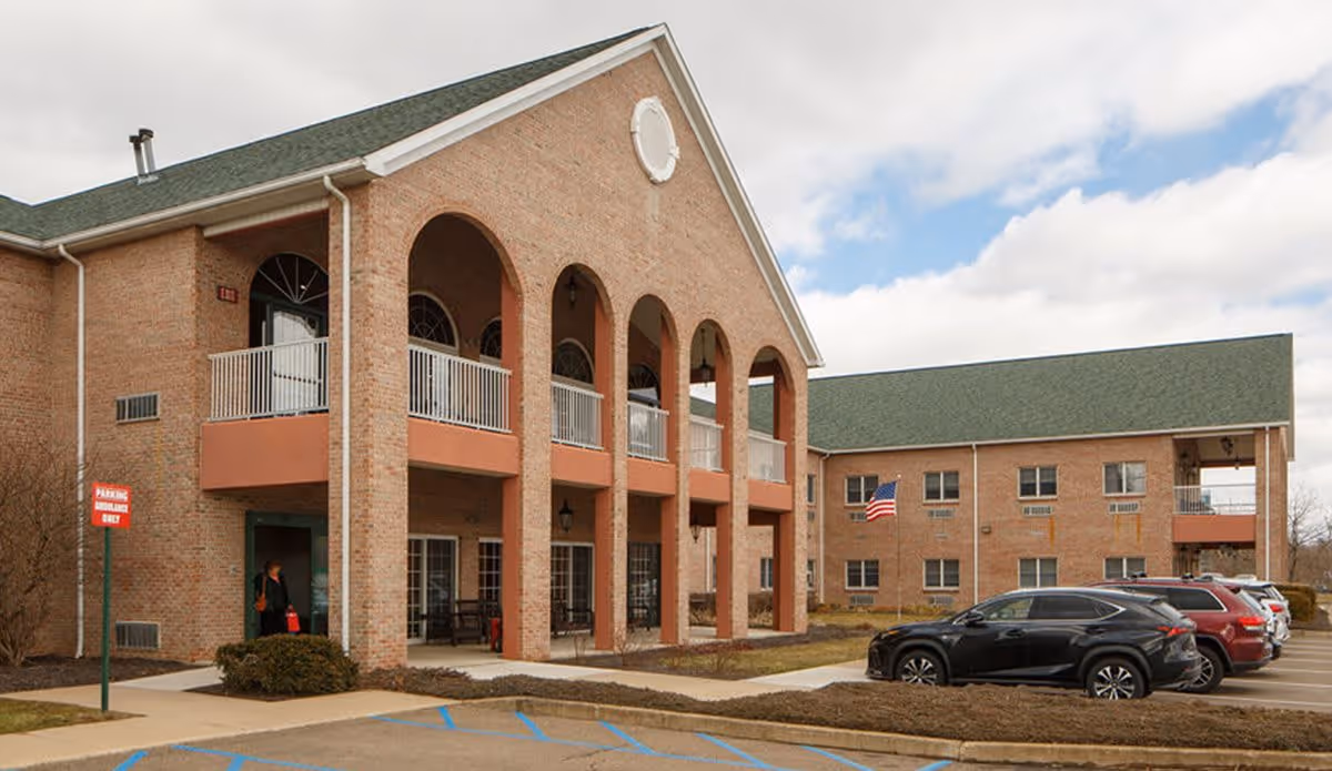 Exterior view of a two-story brick senior living facility with arched balconies, several parked cars, an American flag, and a person walking near the entrance.