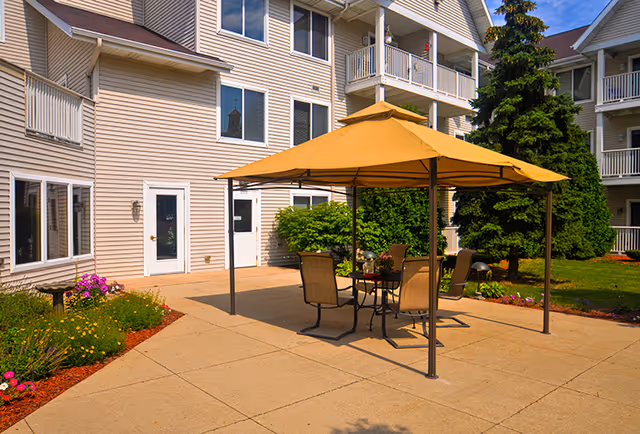 Outdoor patio area at Meadowmere & Mitchell Manor West Allis with a yellow canopy covering a table and four chairs. The patio is surrounded by a garden with flowers and greenery, and the building's exterior with multiple windows and balconies is visible in the background.