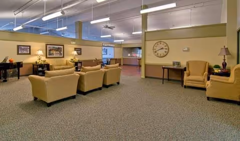 Spacious senior living lounge with sofas and armchairs arranged around side tables and lamps under overhead lighting.