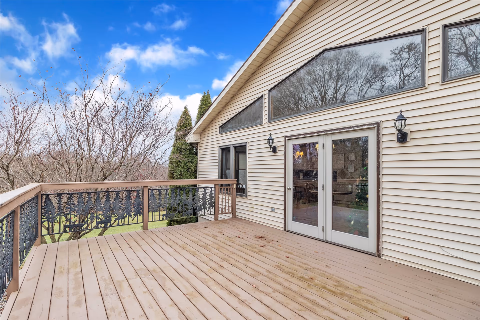 A spacious wooden deck attached to a beige house with large triangular windows and double glass doors. The deck has a decorative metal railing with a nature-inspired design. Leafless trees and a green lawn are visible in the background under a partly cloudy blue sky.