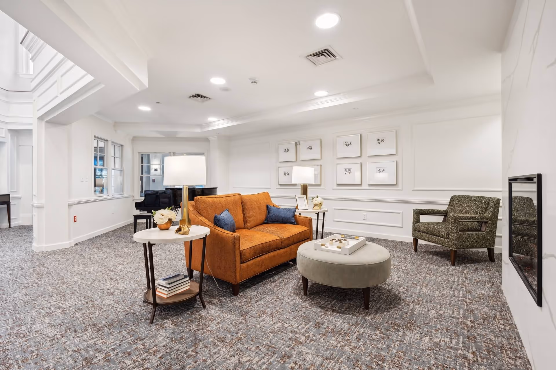 A bright and spacious living room area with a brown loveseat adorned with blue pillows, a round gray ottoman with a game on top, a green armchair, and two side tables with lamps and decorative items. The room has white walls with framed artwork, a carpeted floor, and recessed ceiling lights. A black piano is visible in the background near windows.