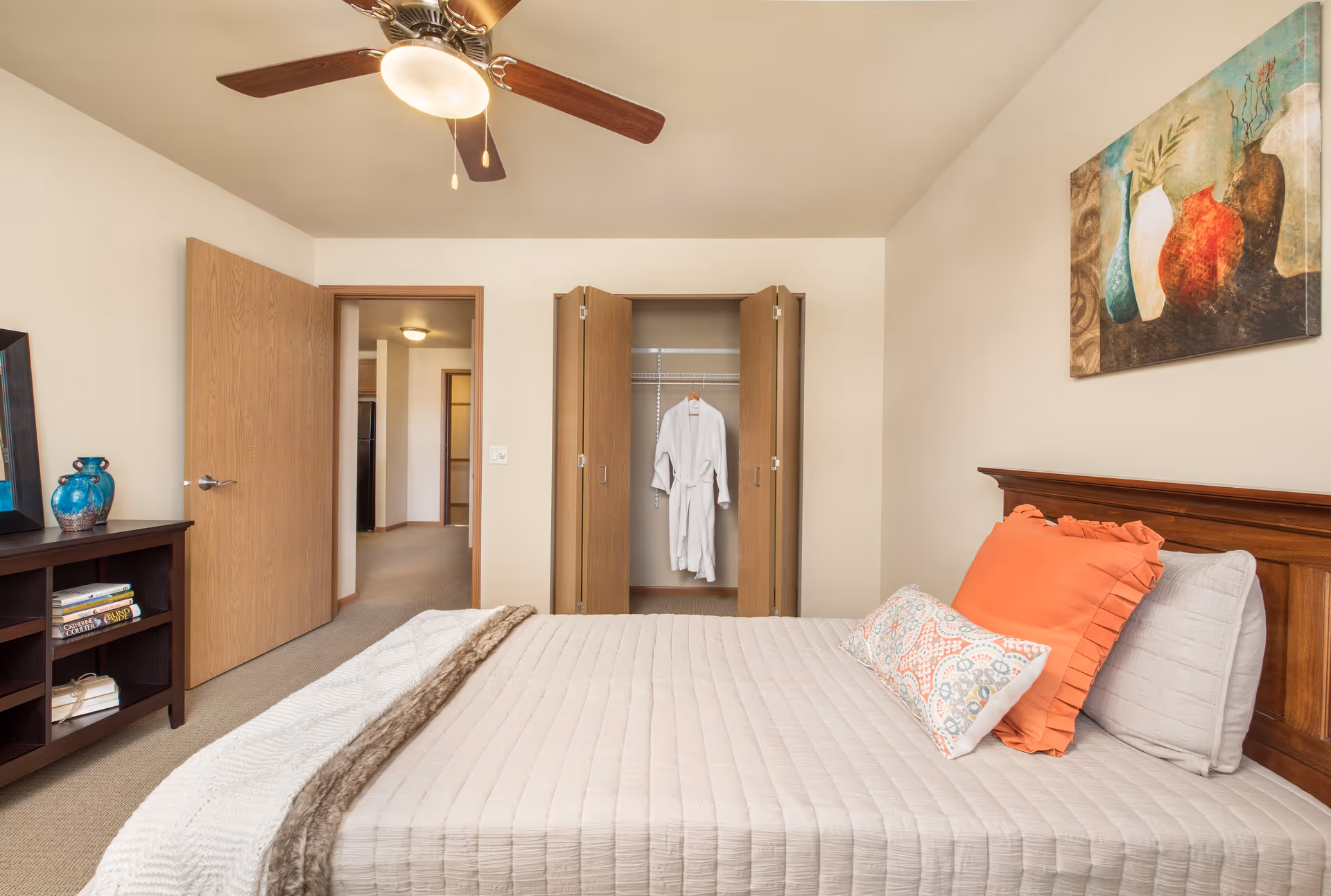 A bedroom with a neatly made bed featuring white and orange pillows and a beige quilt. There is a wooden headboard and a colorful painting of vases on the wall above it. To the left, there is a dark wooden shelf with books and decorative blue vases. The closet doors are open, revealing a white robe hanging inside. A ceiling fan with lights is visible on the ceiling.