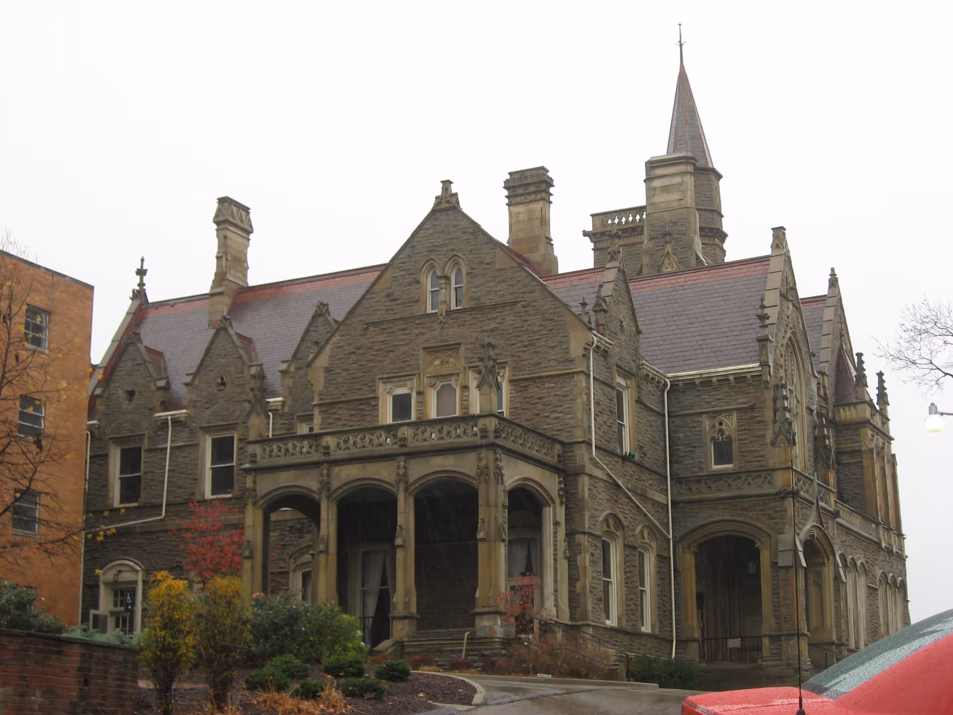 Exterior view of a large, historic stone building with gothic architectural features including pointed arches, decorative stonework, and a tall spire. The building is surrounded by some shrubs and trees, and a red car is partially visible in the foreground.
