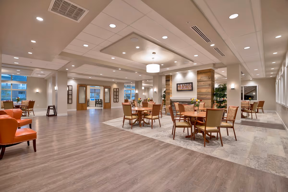 Spacious and well-lit common area in a senior living facility with multiple round tables and chairs arranged for dining or socializing. The room features wood flooring, a section with patterned carpet, modern ceiling lights, and a decorative wall with wood paneling and a framed picture. Large windows and glass doors provide natural light.