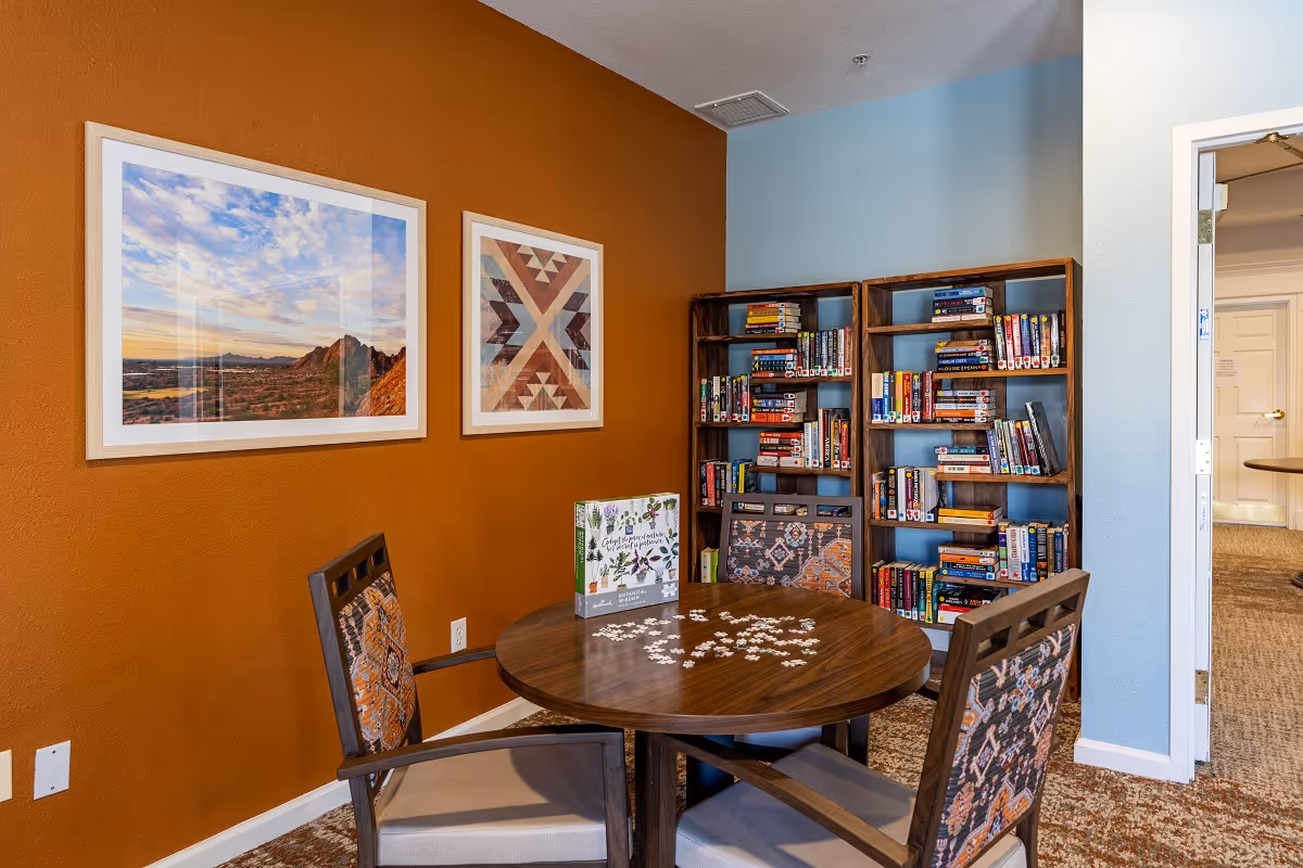 A cozy reading and puzzle area in a senior living facility featuring a round wooden table with a partially completed puzzle and a puzzle box on top. Surrounding the table are four chairs with patterned upholstery. Behind the table are two wooden bookshelves filled with books and DVDs. The walls are painted in warm orange and light blue tones, adorned with two framed pictures, one of a scenic landscape and the other with a geometric pattern. A doorway leads to another carpeted room.
