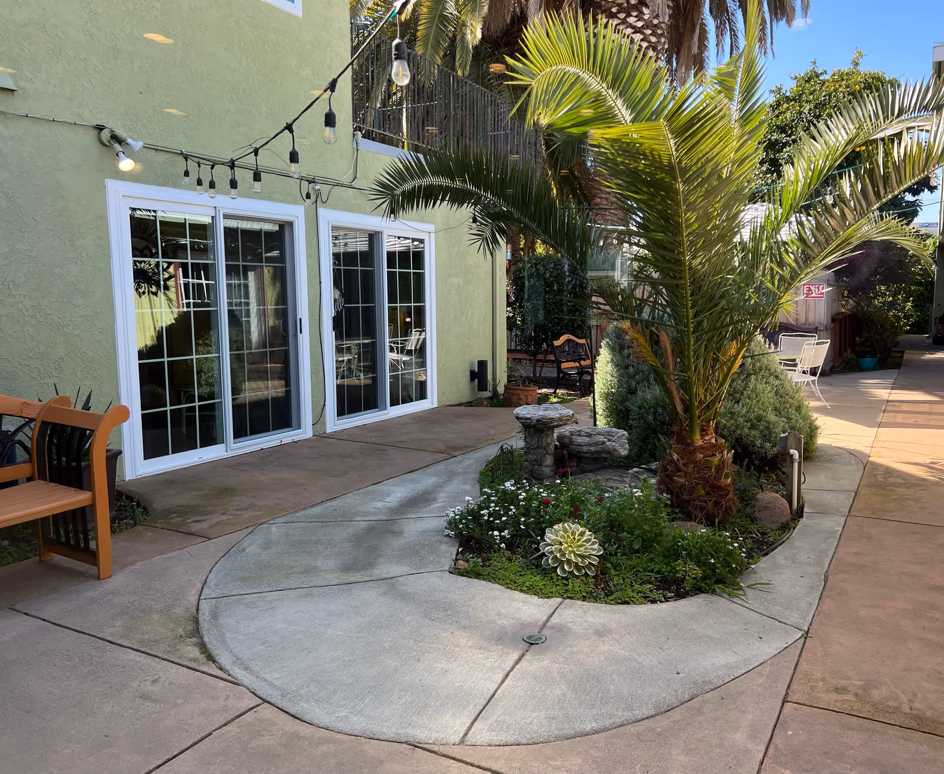 Outdoor patio area at Rosemont Gardens featuring a curved concrete walkway around a landscaped garden bed with a palm tree, small shrubs, and flowers. There are string lights hanging above, a wooden bench on the left, and sliding glass doors leading inside a light green building.