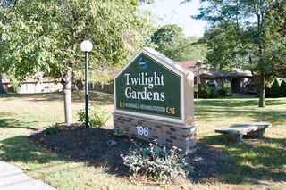 Exterior view of Twilight Gardens Nursing & Rehabilitation with sign, trees, and a bench.