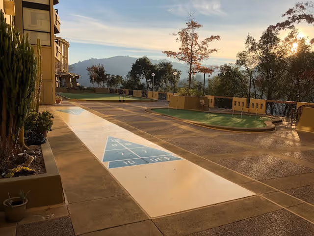 Outdoor recreational area at a senior living facility with a shuffleboard court in the foreground, surrounded by landscaped plants and trees. The setting sun casts a warm glow over the scene, with hills and trees visible in the background.