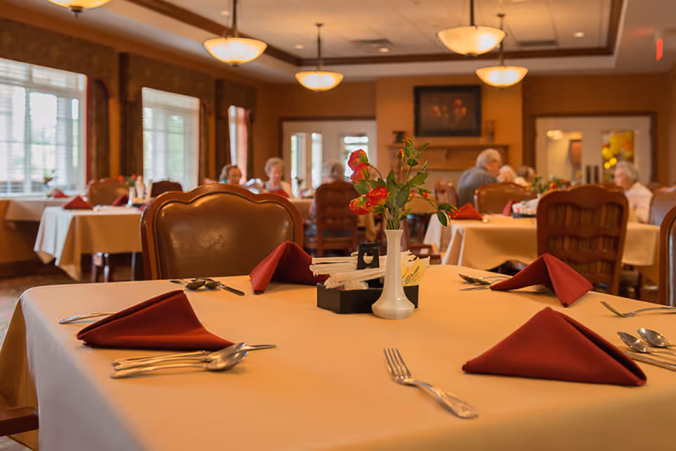 A dining room in a retirement village with tables covered in beige tablecloths, each set with red folded napkins, silverware, and a white vase with red flowers. Several elderly people are seated and conversing in the background. The room has warm lighting and large windows with blinds.