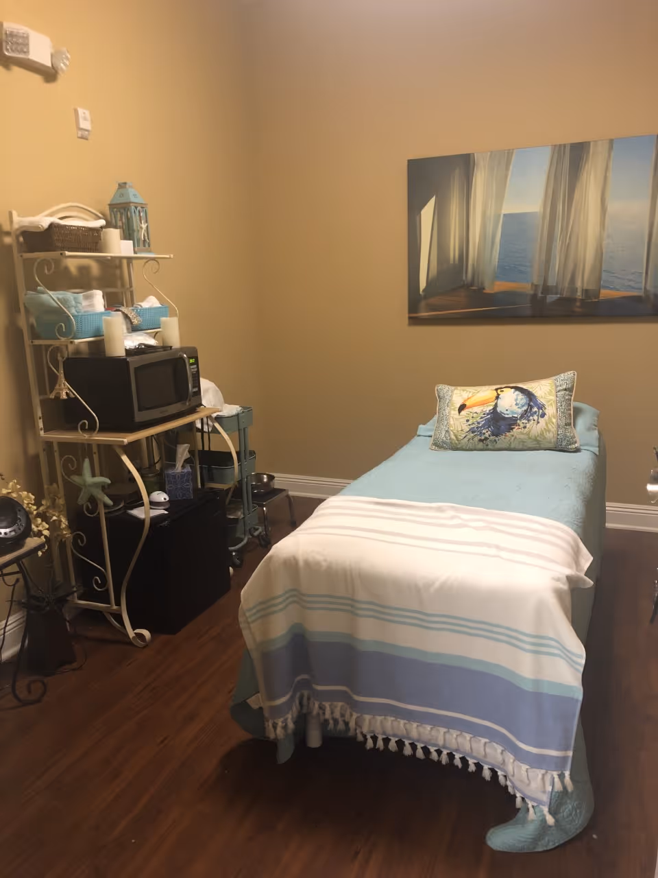 A cozy room with a single bed covered in a light blue quilt and a striped blanket at the foot. There is a decorative pillow with a toucan bird design on the bed. To the left, a white metal shelving unit holds a microwave, baskets, and candles. The floor is wooden, and a large framed picture of a window with sheer curtains overlooking the ocean hangs on the wall.