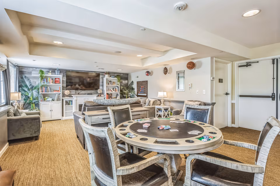 A cozy common area in a memory care facility featuring a round poker table with cards and poker chips, surrounded by four chairs. In the background, there are comfortable sofas, a large flat-screen TV mounted on a wall with built-in white bookshelves on either side, and decorative plants. The room has beige walls, carpeted flooring, and recessed ceiling lights.