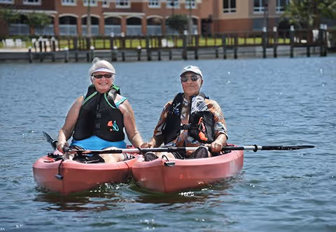 Two elderly people wearing life jackets and sunglasses are sitting in a red tandem kayak on a body of water with buildings and greenery in the background.