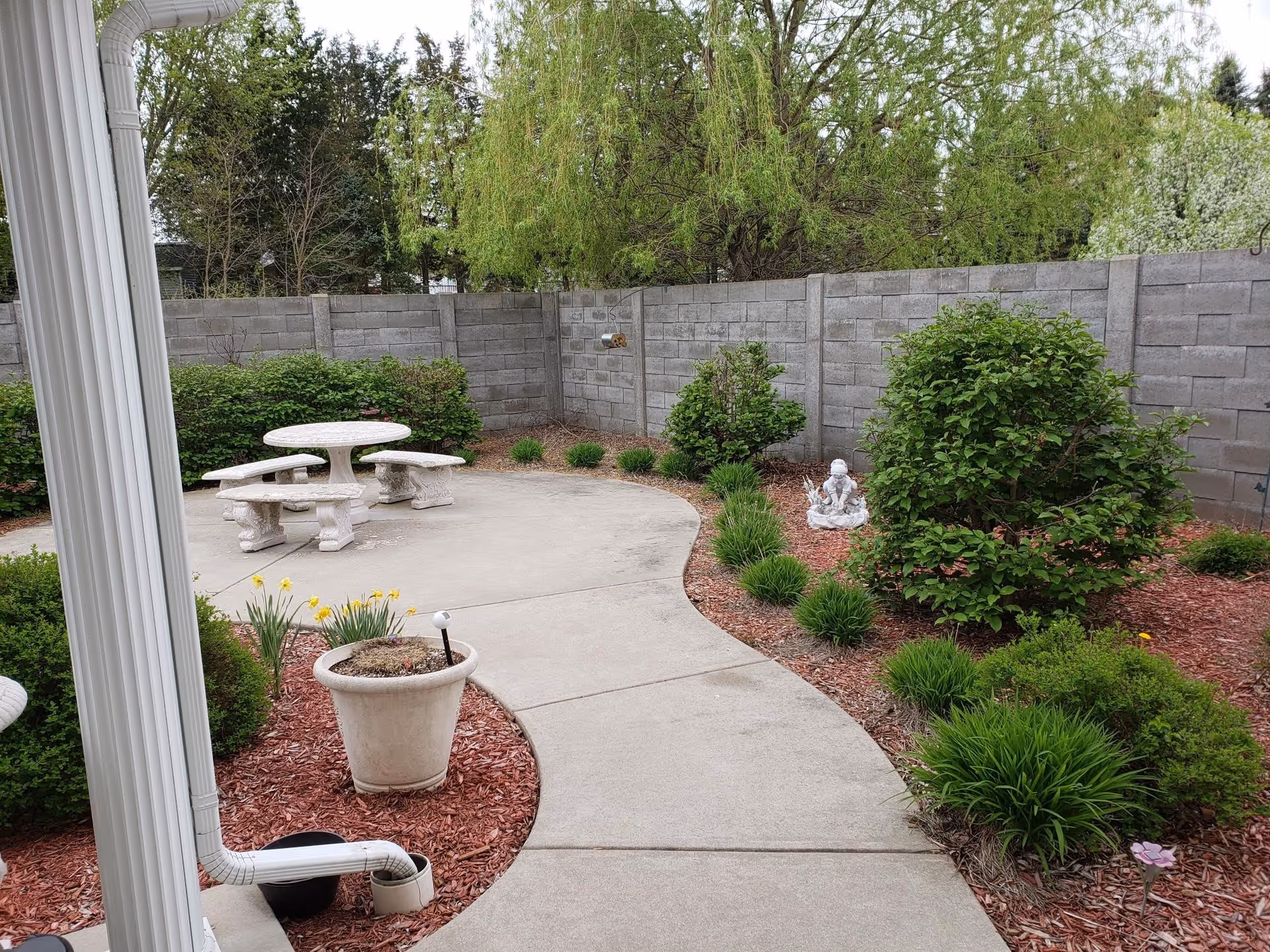 Outdoor garden area with a curved concrete pathway leading to a round stone table with matching benches. The garden is bordered by a gray concrete block wall and features various green bushes, plants, and a small statue of a cherub. There is a white downspout on the left side and a large planter with some flowers near the pathway.