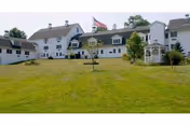 Large white multi-story building with multiple windows and a gray roof, situated on a grassy hill with a flagpole flying the American flag and a white gazebo to the right.