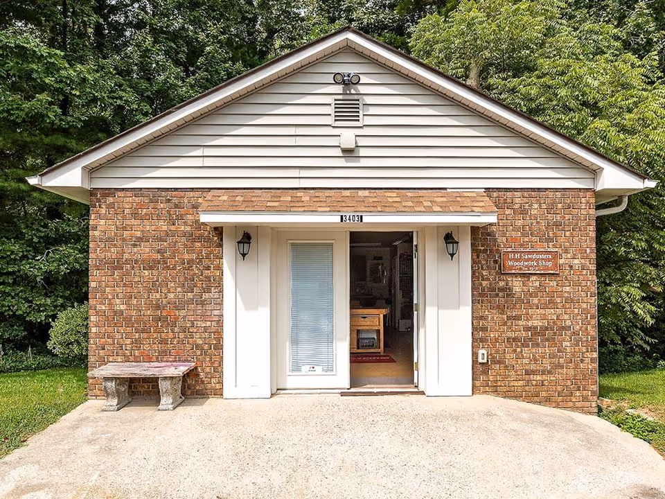 Front exterior view of a small brick building with a white door and a sign that reads 'H H Sawdusters Woodwork Shop'. There is a concrete bench to the left of the door and trees surrounding the building.