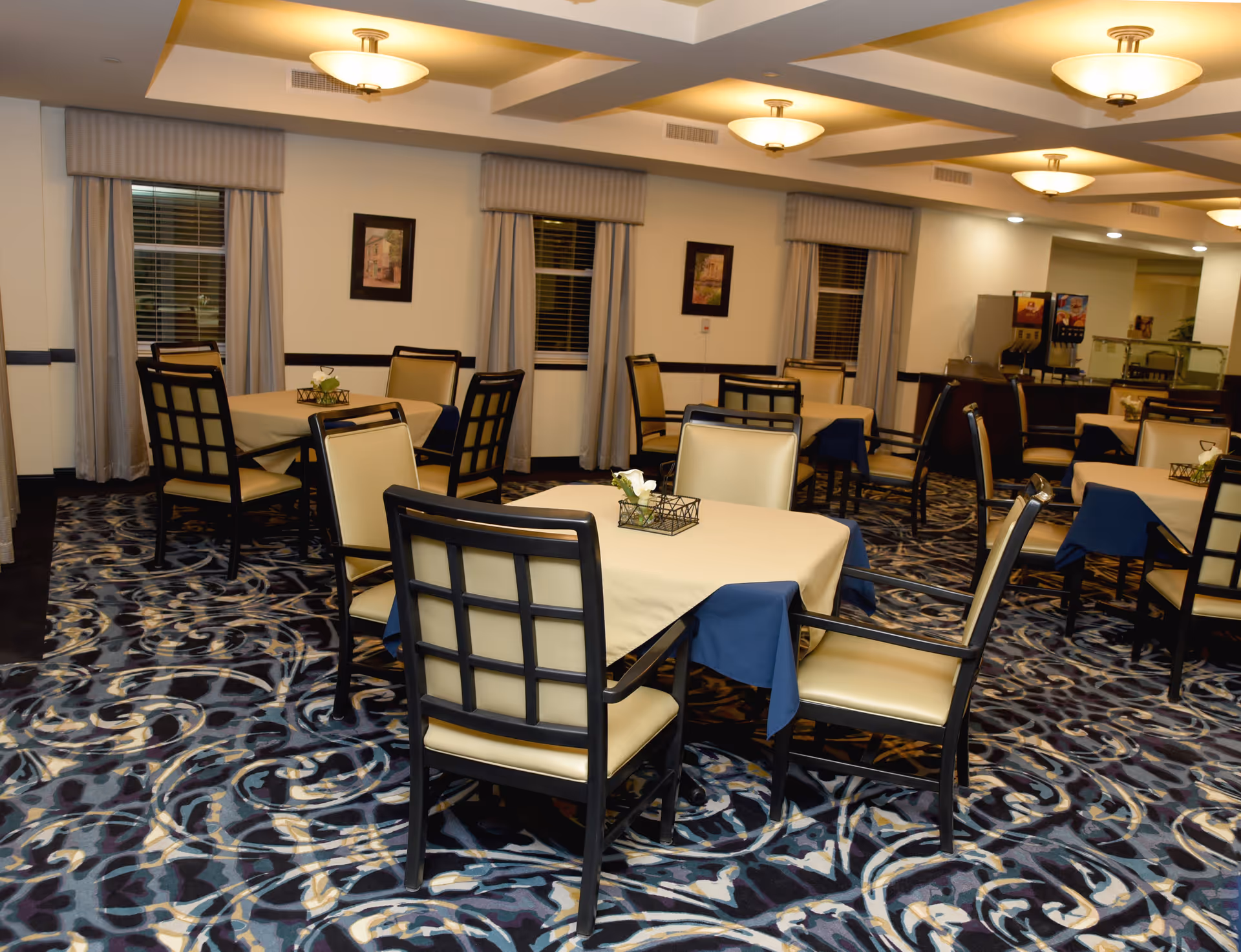 Interior view of a dining room with multiple tables covered with beige and blue tablecloths, surrounded by cushioned chairs with black frames. The room has patterned carpet flooring, framed pictures on the walls, windows with curtains, and ceiling lights providing warm illumination. A beverage dispenser is visible in the background.