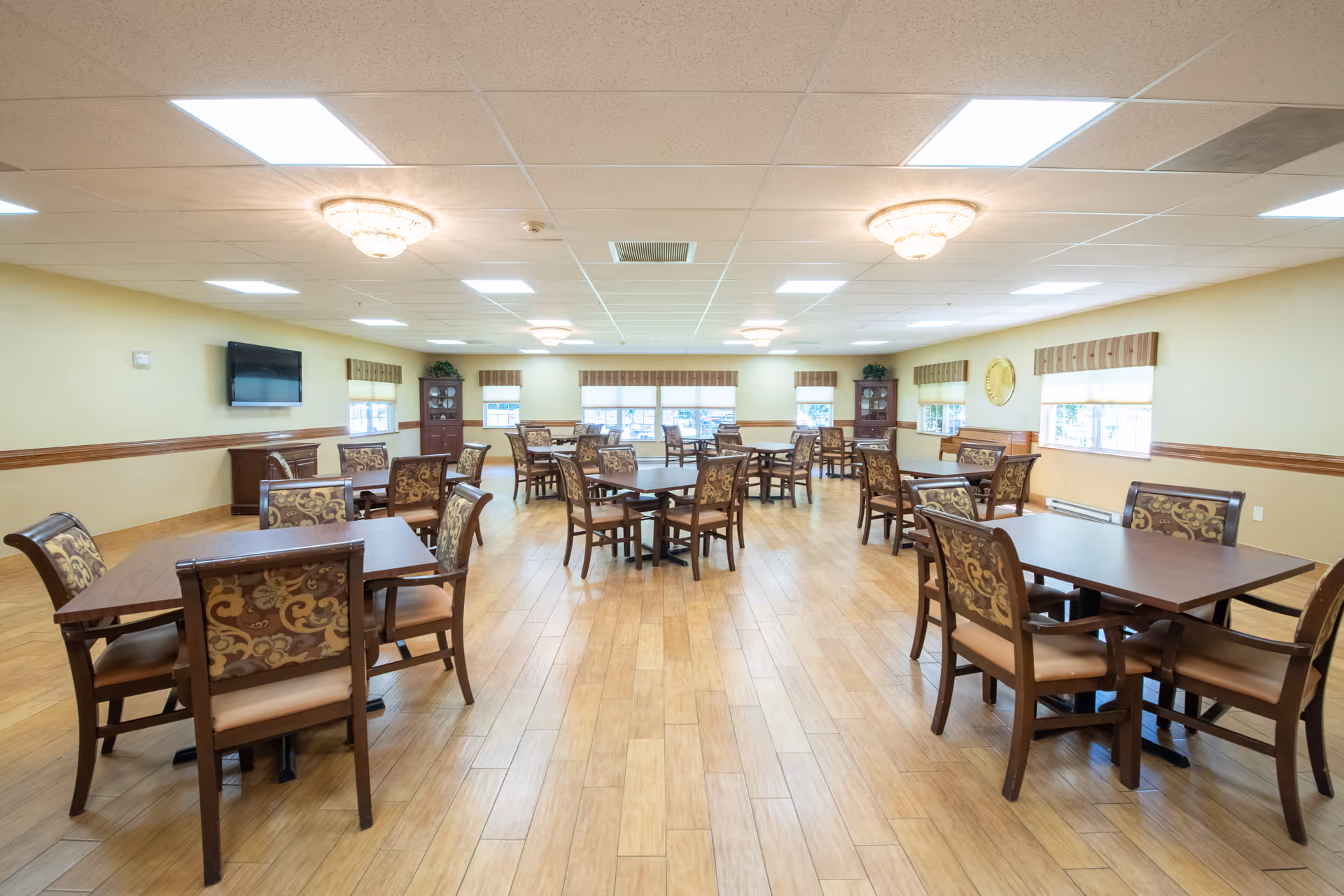 A spacious dining room with multiple wooden tables and chairs arranged neatly. The chairs have patterned upholstery. The room has light-colored walls, wooden flooring, several windows with valances, and ceiling lights including chandeliers. A flat-screen TV is mounted on one wall, and there are cabinets and decorative plants in the corners.