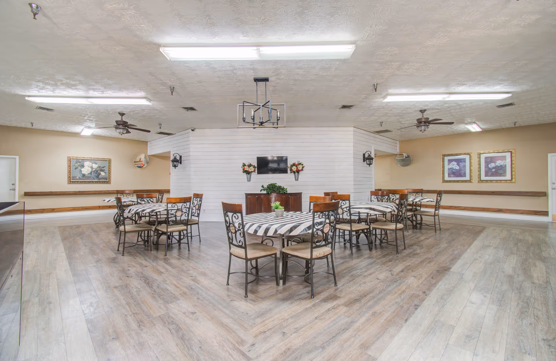 A spacious dining room with several round tables covered with striped tablecloths and surrounded by chairs. The room has light wood flooring, beige walls, ceiling fans, and fluorescent lighting. There are framed floral paintings on the walls and a small cabinet with a TV and flower arrangements in the center background.