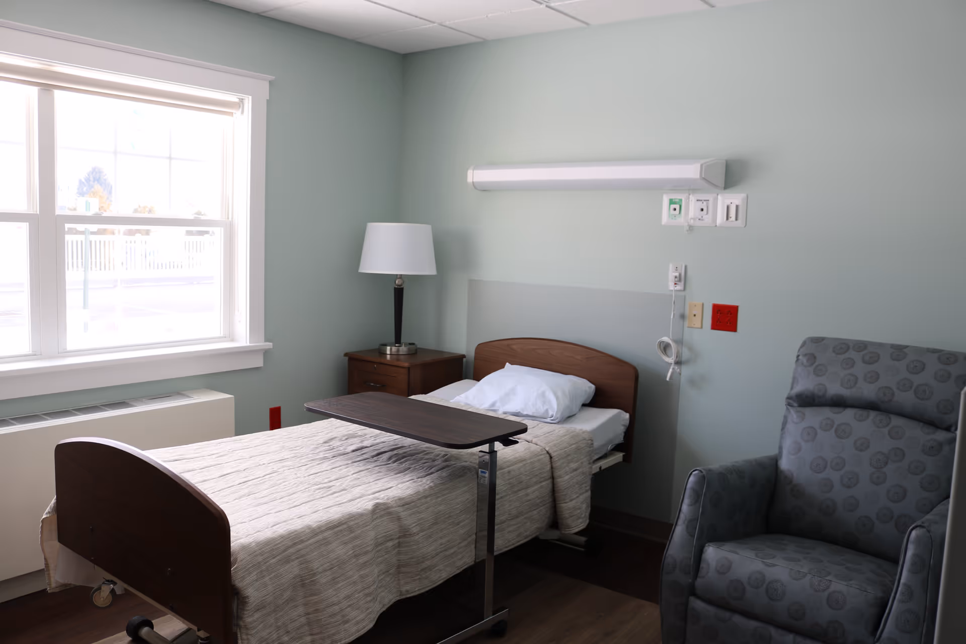 A small, clean personal care room with a single hospital-style bed covered with a beige blanket and a white pillow. Next to the bed is a wooden nightstand with a lamp. A gray patterned recliner chair is positioned near the bed. The room has light green walls, a large window letting in natural light, and medical equipment mounted on the wall above the bed.