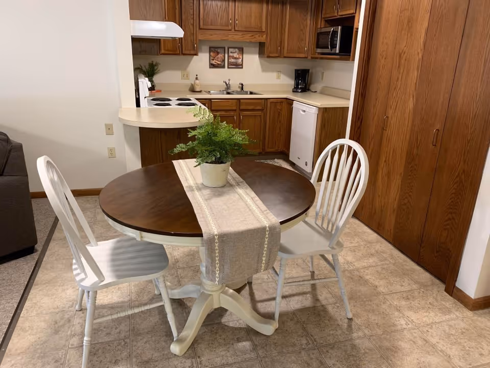 Small dining area with a round wooden table and two white chairs in front of a compact kitchen with wood cabinets.