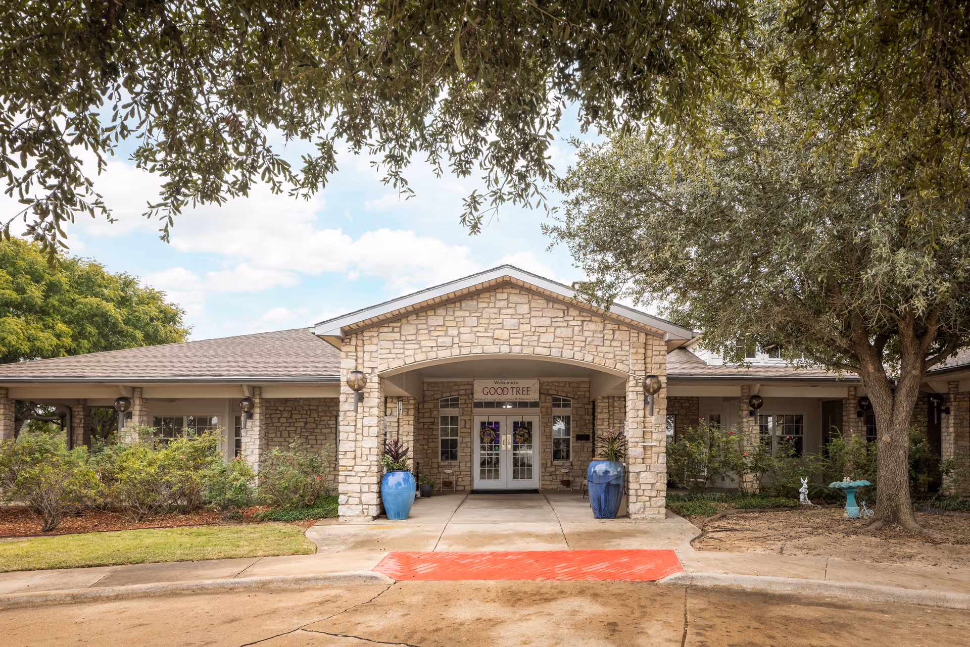 Front exterior view of a single-story stone building with a covered entrance. Two large blue planters with plants flank the double glass doors under an archway. Trees and shrubs surround the building, and a red painted curb is visible in front.