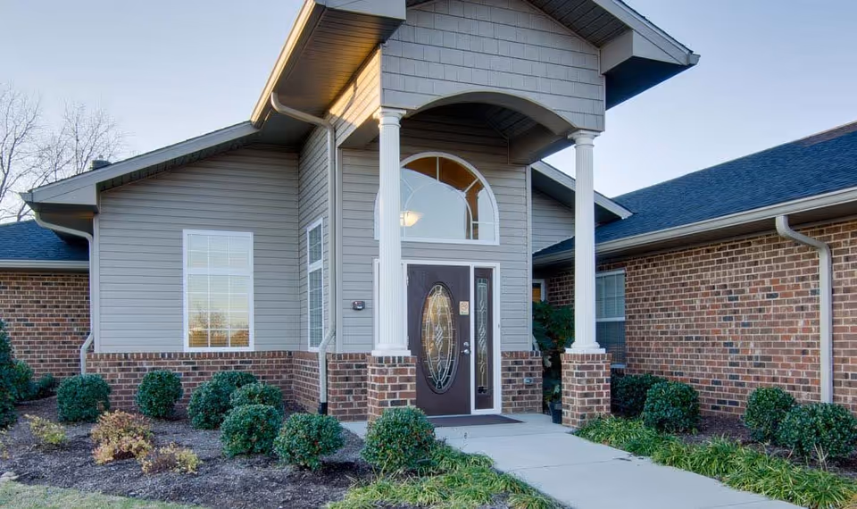 Front entrance of a senior living facility with a covered porch supported by white columns, a decorative glass front door, brick and siding exterior walls, and neatly trimmed bushes along the walkway.