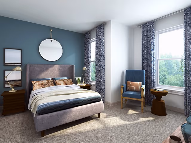 A bright and cozy bedroom with a large upholstered bed featuring patterned pillows and a soft blanket. The room has a blue accent wall with a round mirror and two framed pictures. There are two bedside tables with lamps and a vase of flowers. Two large windows with patterned curtains let in natural light, and a blue armchair with a cushion sits next to a small wooden side table.