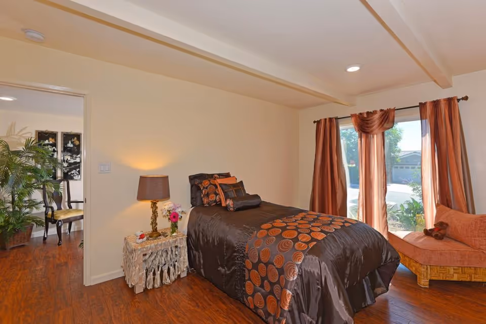 A cozy bedroom with a single bed covered in dark brown and orange bedding. Next to the bed is a small table with a lamp and a vase of flowers. There is a window with sheer and solid orange curtains letting in natural light. A cushioned bench is placed near the window. The floor is wooden, and an open doorway leads to another room with chairs and a large plant.