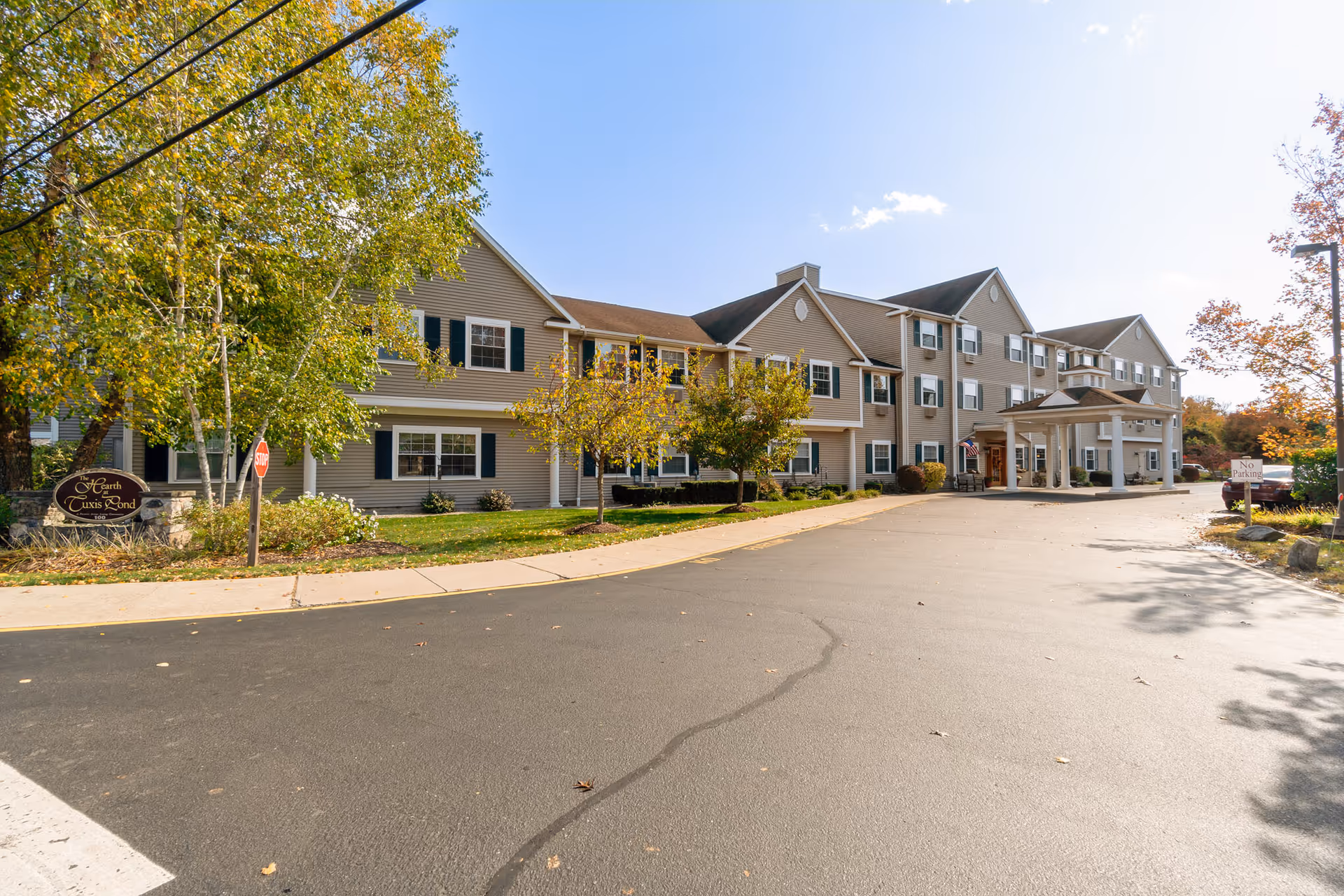 Exterior view of a multi-story senior living facility building with beige siding, white trim, and green shutters. The building has a covered entrance with columns and is surrounded by trees with autumn foliage. A paved driveway and sidewalk lead to the entrance. A sign near the building reads 'The Hearth at Tuxis Pond.'