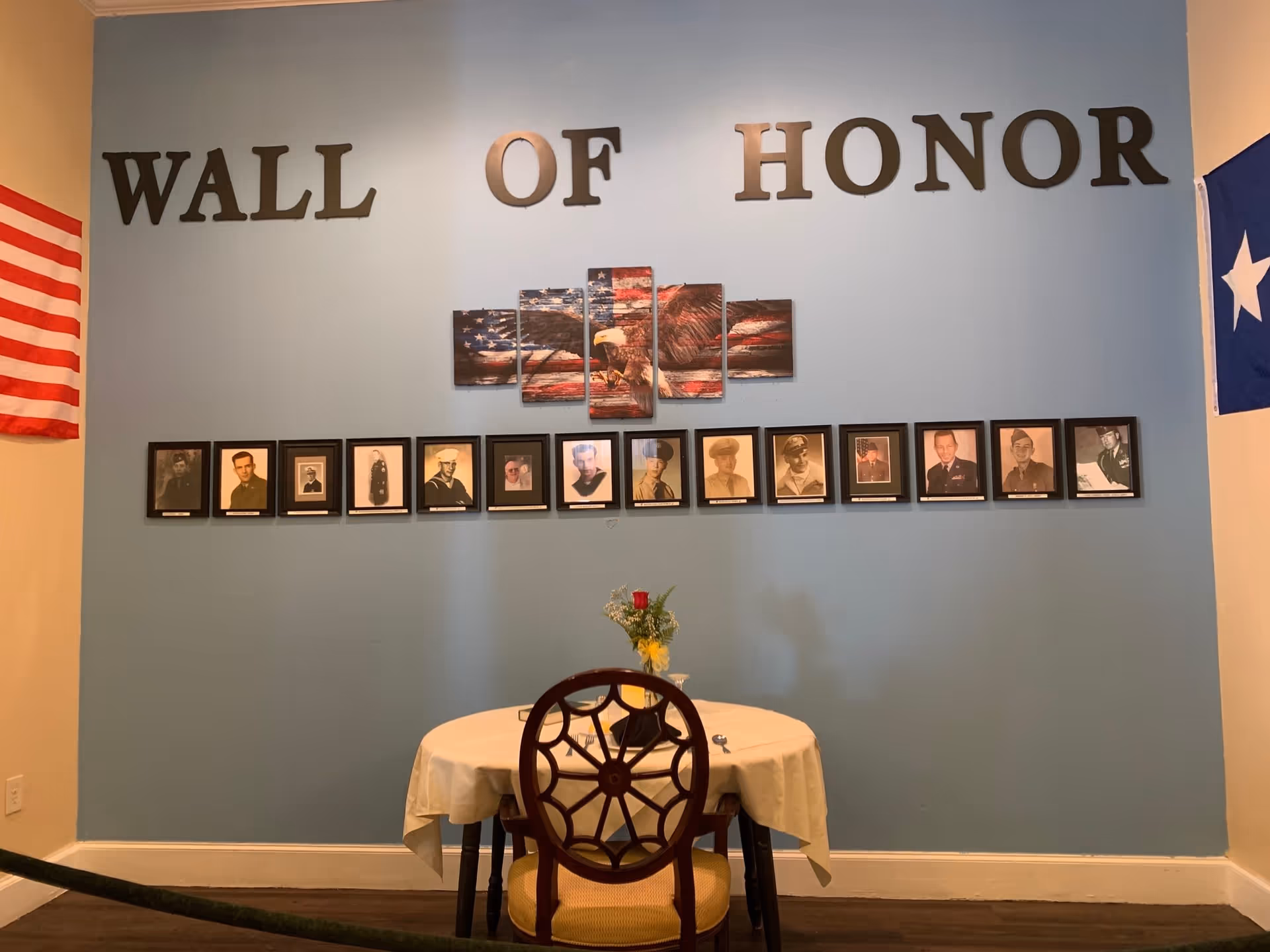 A Wall of Honor display with framed photographs of veterans arranged in a row on a light blue wall. Above the photos, large black letters spell out 'WALL OF HONOR'. In the center above the photos is a multi-panel artwork of a bald eagle with an American flag background. Below the display is a small round table covered with a white tablecloth, set with a single chair and a small floral arrangement with a red rose. American flags are partially visible on the left and right edges of the image.