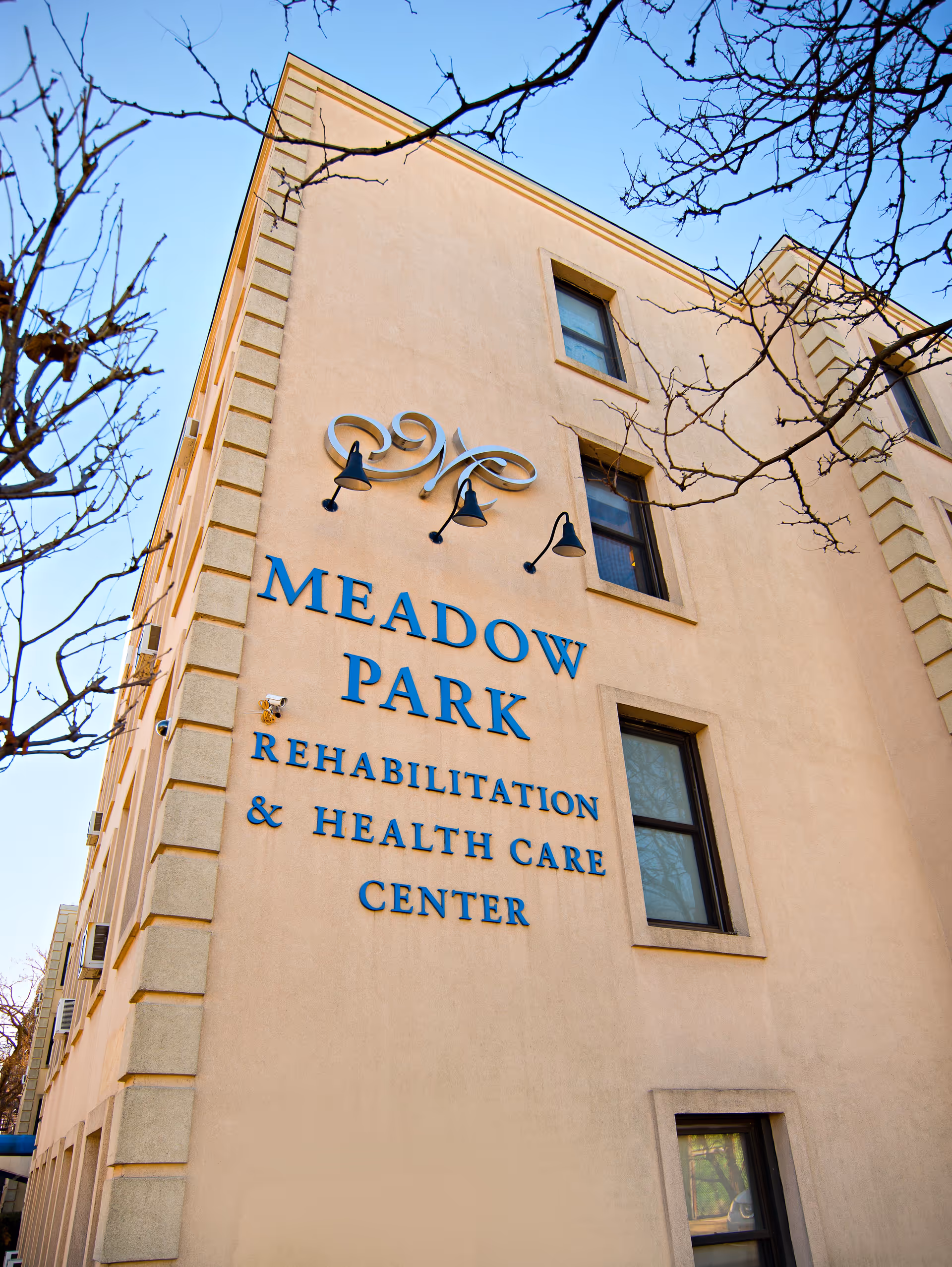 Exterior view of a beige multi-story building with the sign 'Meadow Park Rehabilitation & Health Care Center' mounted on the wall. There are three windows visible and some leafless tree branches in the foreground against a clear blue sky.