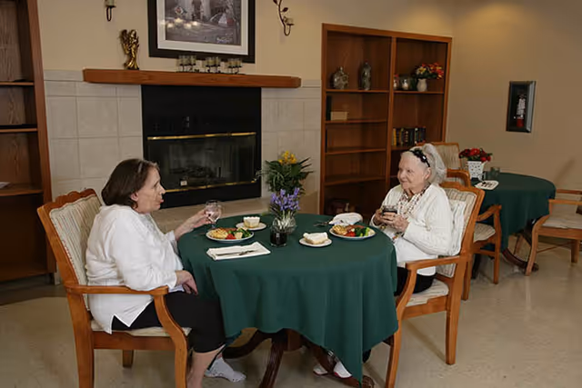 Two elderly women sitting at a round table with a green tablecloth, having a meal and drinks in a cozy room with a fireplace, wooden bookshelves, and a framed picture above the fireplace.