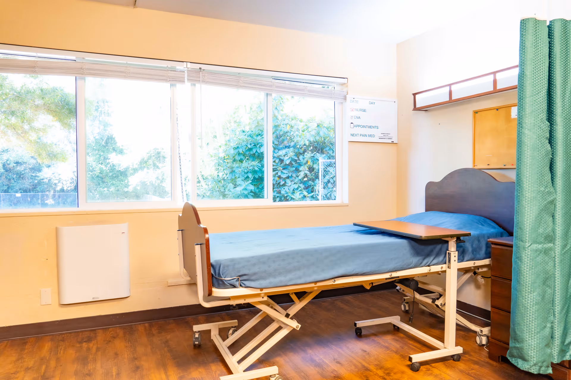 A hospital-style bed with a blue mattress and an adjustable overbed table in a room with large windows showing greenery outside. There is a green privacy curtain on the right side and a whiteboard on the wall with notes about nurse, CNA, appointments, and next pain medication.