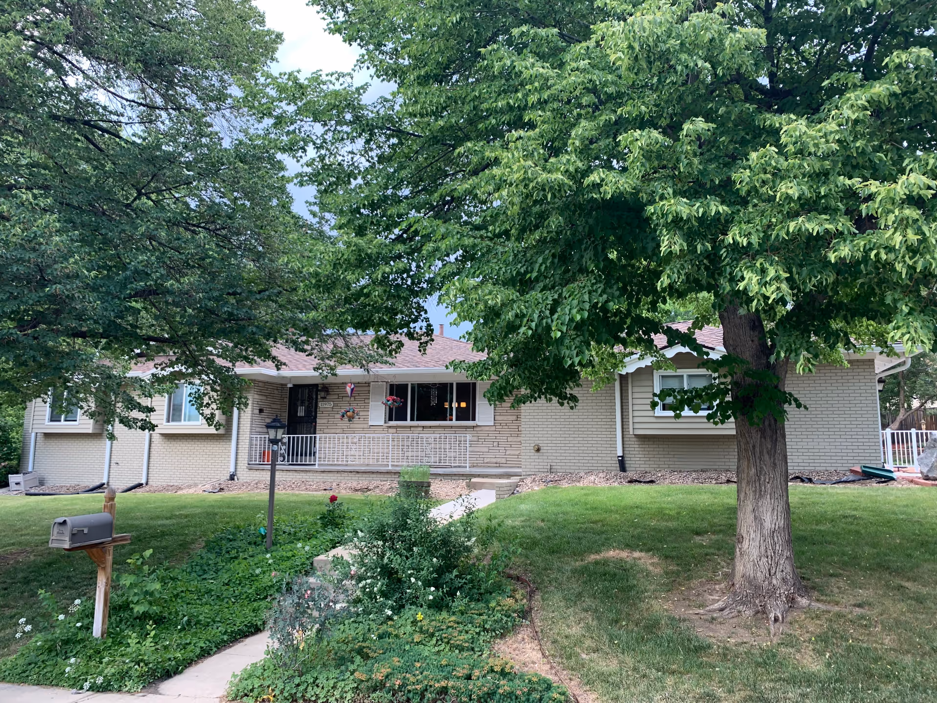 Single-story brick house with a front porch, surrounded by green trees and a well-maintained lawn. A mailbox is visible on the left side near the sidewalk leading to the porch.