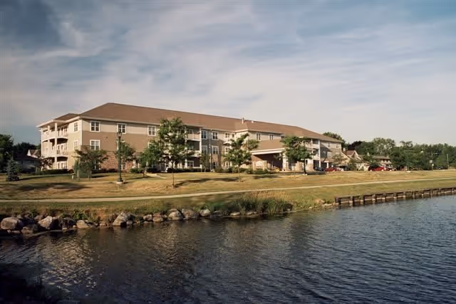 A large, multi-story residential building with balconies overlooking a calm body of water. The building is surrounded by a grassy area with trees and a clear sky above.