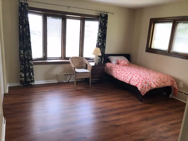 A simple bedroom with a single bed covered in a pink and white patterned bedspread, a wooden nightstand with a lamp, a wicker chair with a cushion, and large windows with dark floral curtains letting in natural light. The room has wooden flooring and beige walls.