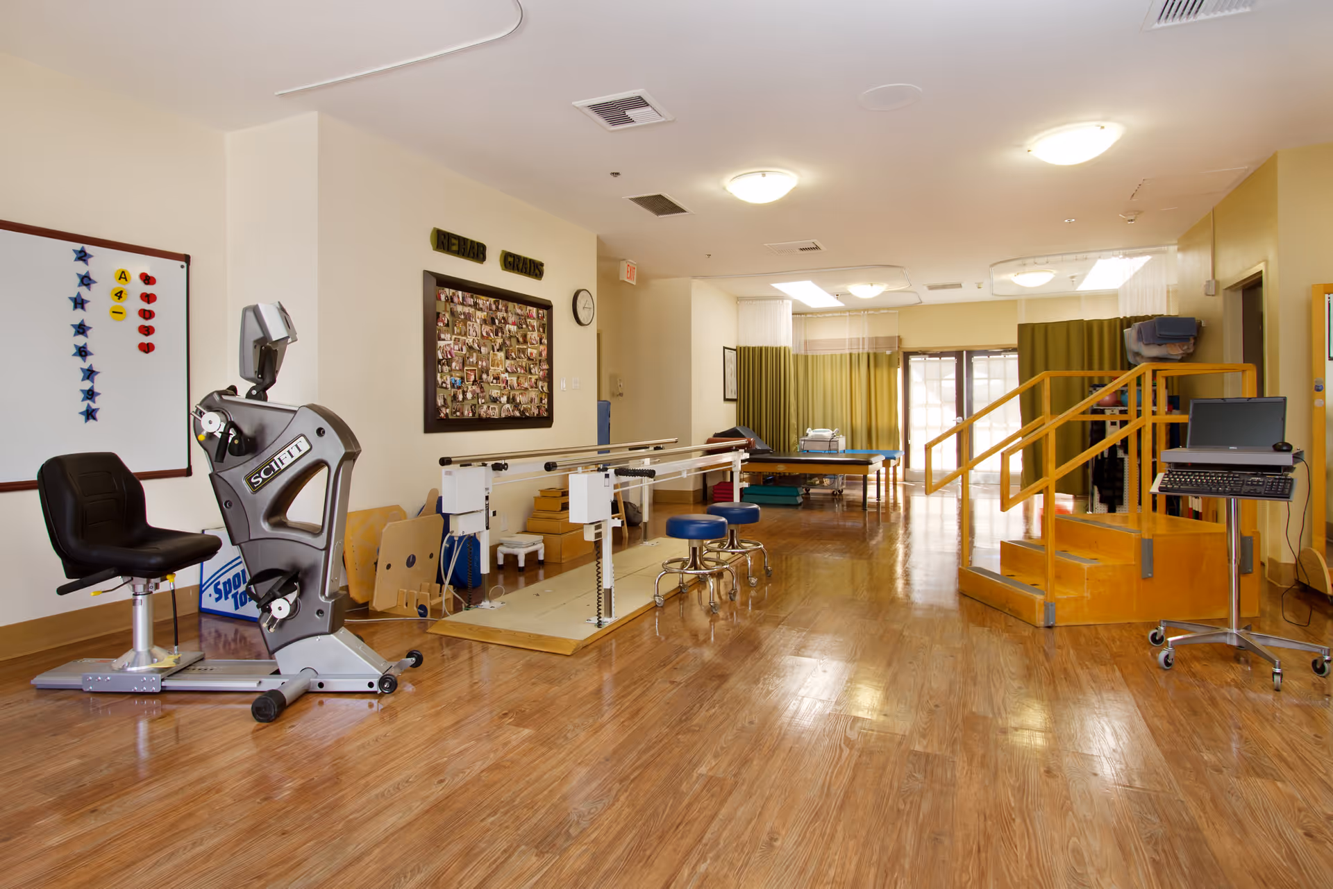 A rehabilitation therapy room with exercise equipment including a stationary bike, parallel bars for walking practice, stools, a set of stairs with handrails for physical therapy, and a treatment table in the background. The room has wooden flooring, bright lighting, and a bulletin board labeled 'REHAB GRADS' with photos on the wall.
