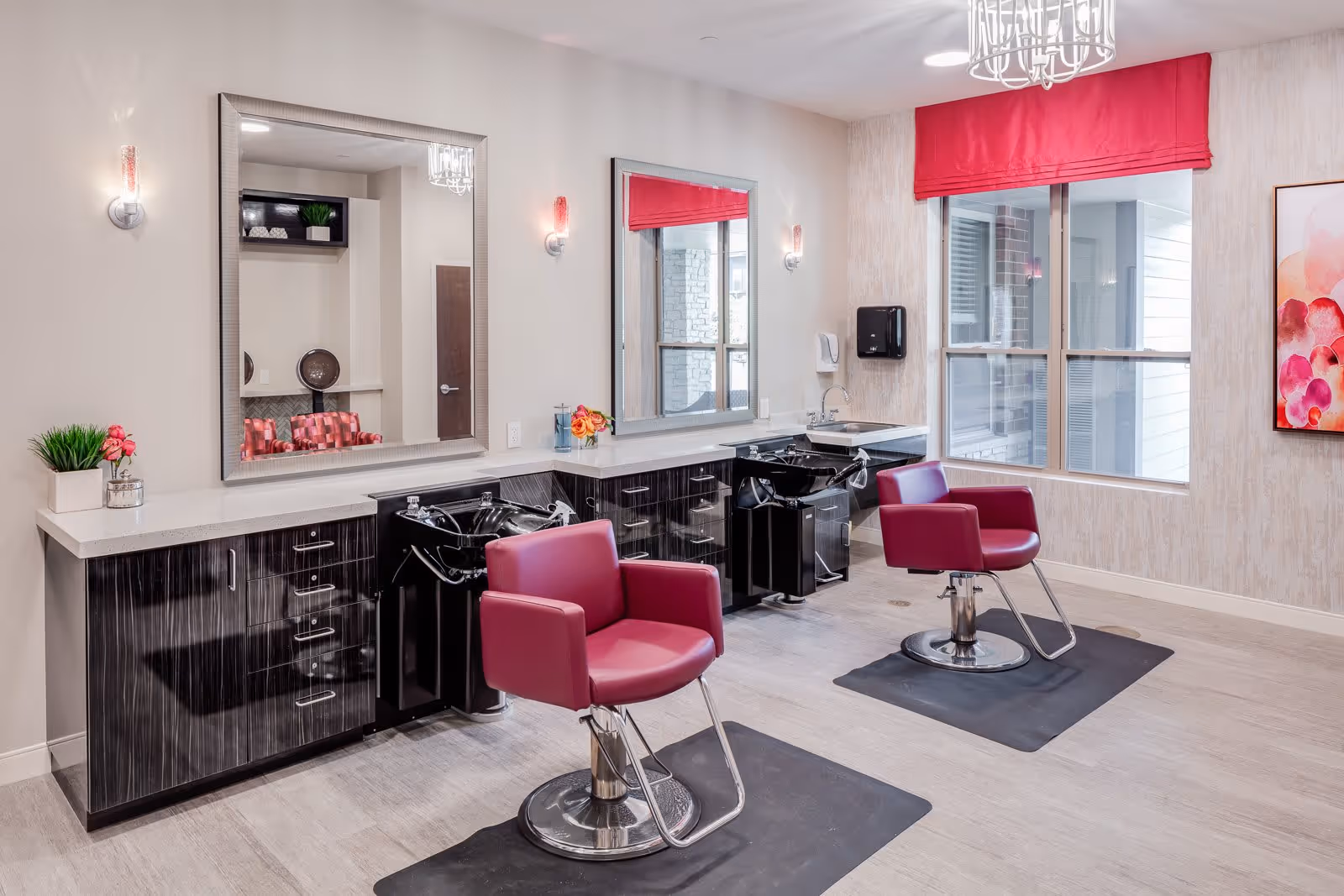 A bright and modern salon area with two red salon chairs positioned in front of black wash basins and large mirrors. The room features light-colored walls, a window with a red valance, decorative wall sconces, a chandelier, and a colorful abstract painting on the wall.