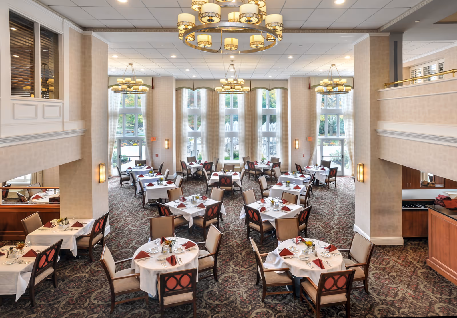 Spacious elegant dining room with multiple round and rectangular tables set with white linens and red napkins, chandeliers, and tall windows.